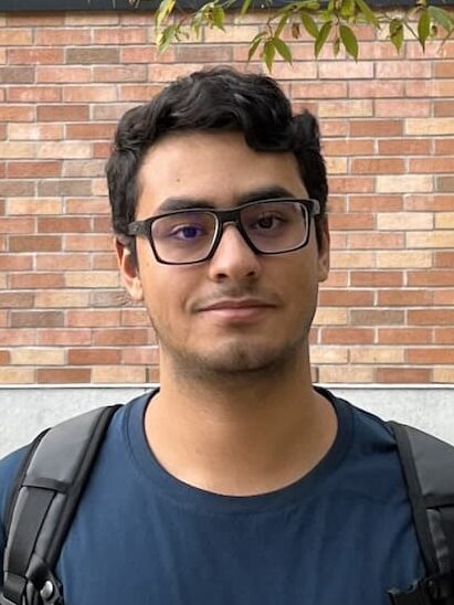 Anas Awadalla, wearing black glasses and a navy t-shirt and a backpack, stands in front of the Paul G. Allen Center for Computer Science & Engineering. The building is made of brick and the sign is black. 