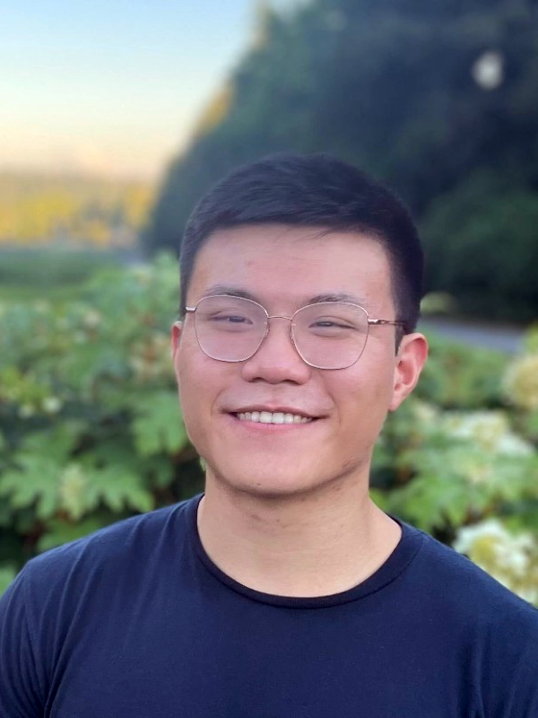 Michael Duan, wearing square glasses and a navy t-shirt, smiles in front of a blurred background of green plants and pine trees. 