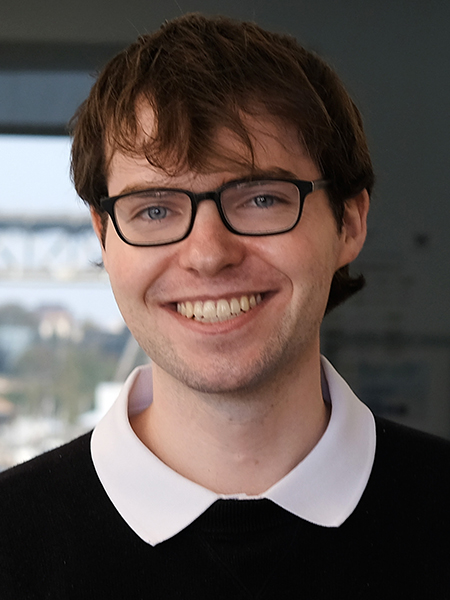 Matt Deitke, wearing glasses and black sweater over a white collared shirt, smiles in front of a blurred background. A window depicting a cityscape is to the left. 