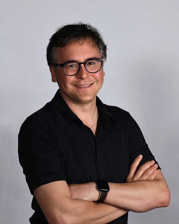 Portrait of Luis Ceze standing with arms crossed, smiling at the camera, against a grey background. Luis is wearing glasses with dark acrylic frames and clear lenses, black short-sleeved, open-necked shirt, and a black smartwatch on his left wrist.