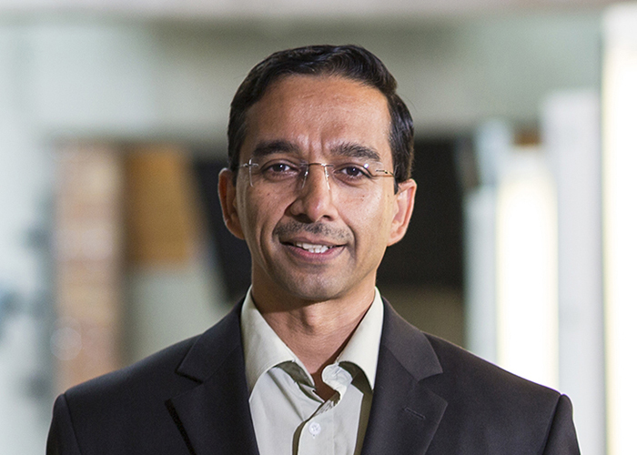Portrait of a smiling Rajesh Rao wearing wire-rimmed eyeglasses and a dark grey suit jacket over a pale grey button-up shirt, with concrete and brick features and catwalk lighting in the Paul G. Allen Center atrium visible in the background.