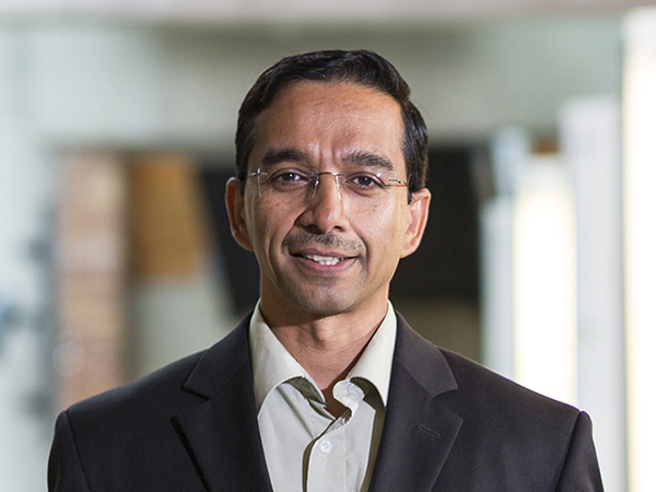 Portrait of a smiling Rajesh Rao wearing wire-rimmed eyeglasses and a dark grey suit jacket over a pale grey button-up shirt, with concrete and brick features and catwalk lighting in the Paul G. Allen Center atrium visible in the background.