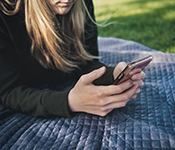 A person with long blond hair, with only mouth and chin visible, is lying on a blue quilted blanket on short green grass in dappled sunlight. The person is wearing a black sweatshirt and propped up on their elbows, viewing a smartphone held in their well-manicured hands.