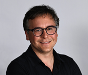 Portrait of Luis Ceze smiling at the camera. Luis is wearing glasses with dark acrylic frames and clear lenses and a black open-necked shirt against a grey background.
