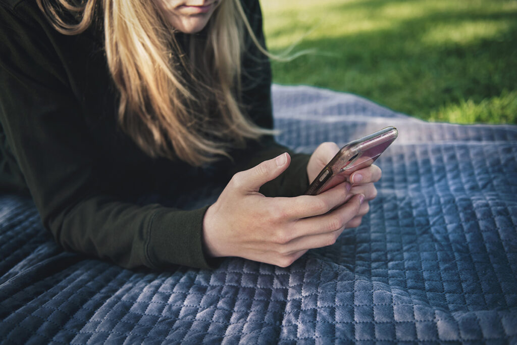 A person with long blond hair, with only mouth and chin visible, is lying on a blue quilted blanket on short green grass in dappled sunlight. The person is wearing a black sweatshirt and propped up on their elbows, viewing a smartphone held in their well-manicured hands.