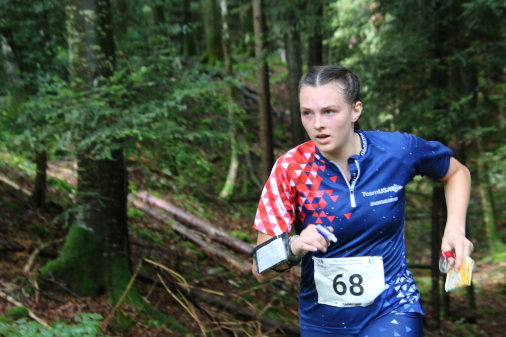 Jessica Colleran, wearing a red and blue Team USA jersey with a triangle pattern on the sleeve and a number 68 on the front, runs through a forest while holding a marker, compass and map and wearing a wristband. 