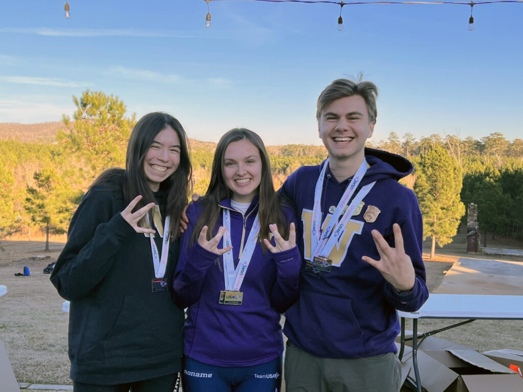 Annika Mihata, wearing a black University of Washington hooded sweatshirt and a medal around her neck, smiles with Jessica Colleran, wearing a purple University of Washington hooded sweatshirt and a medal, and Curtis Anderson, wearing a purple University of Washington hooded sweatshirt and a medal. They are making W signs with their hands and are standing in front of a wooded background. 