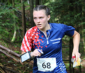 Jessica Colleran, wearing a red and blue Team USA jersey with a triangle pattern on the sleeve and a number 68 on the front, runs through a forest while holding a marker, compass and map and wearing a wristband.