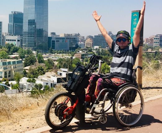 Felipe Tapia, a project participant in Latin America, rides trails on an adapted bicycle and collects sidewalks and path data in his city, Santiago in Chile. He is wearing sunglasses, a ballcap, a striped black and grey t-shirt. He is on a hill overlooking the city. 