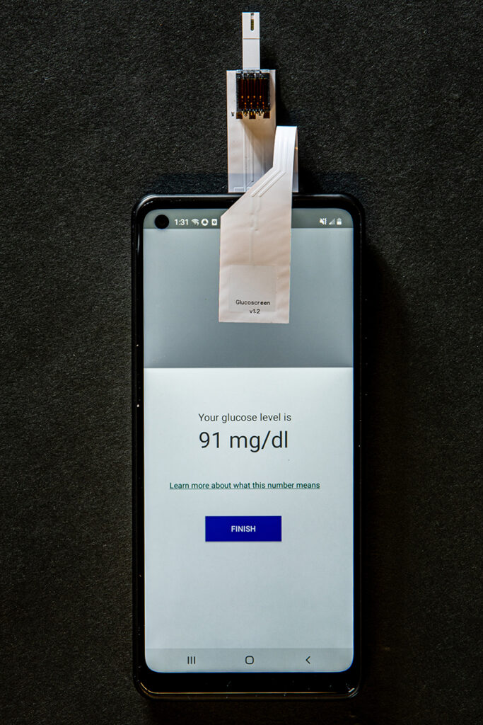 A smartphone with a glucose test strip affixed to the front and rear, with a biosensor and strip for applying a drop of blood sticking out above the phone's top edge. The phone's touch screen is displayed, with the end of the test strip that comes up over the top edge of the phone affixed to the upper third of the screen, which is blank except for a pale grey. The rest of the screen is white with text: Your glucose level is 91 mg/dl, a text link: Learn more about what this number means, and a blue button labeled: Finish.