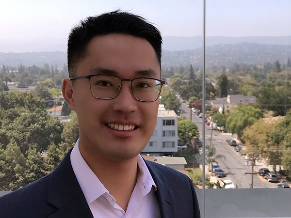 Portrait of Sheng Wang wearing glasses and a navy blue suit jacket over a pink button-down shirt, standing in front of a window on a high floor overlooking low-rise buildings surrounded by trees with a mountain range barely visible in the background.