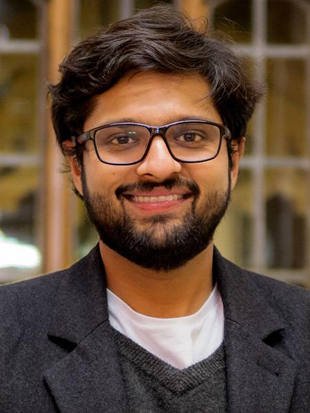 Portrait of Dhruv Jain smiling and wearing dark framed acrylic glasses, white t-shirt, charcoal v-neck sweater, and charcoal wool blazer standing in front of a grid window of leaded glass framed by wood.