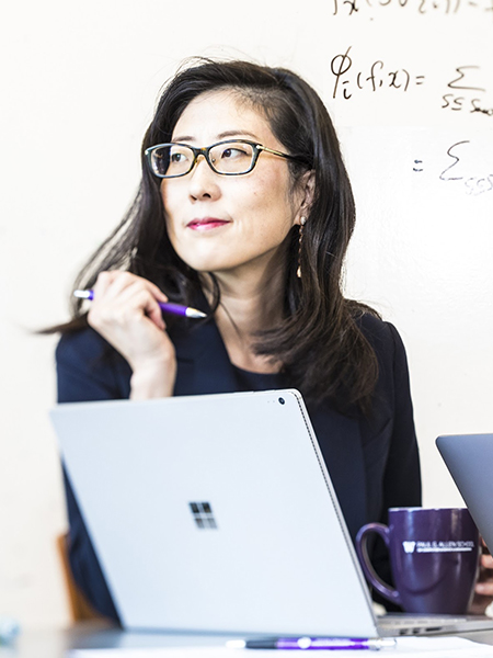 Portrait of Su-In Lee seated at a table in front of a white board, wearing glasses and a black suit and looking off to the viewer's left (her right), holding a pen in her right hand with her elbow on the table and her left hand around a purple and white mug. A second pen and paper is visible lying flat on the table in front of the open laptop, and the corner of a second laptop is just visible in the right of the frame.