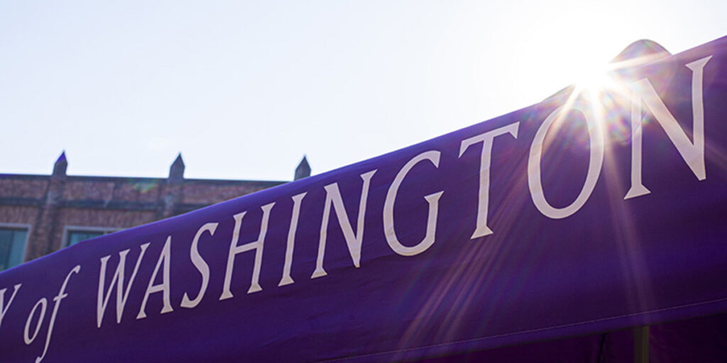 The word "Washington" in University of Washington font in white on a purple fabric banner mostly obscuring campus buildings, backed by a pale blue sky and a burst of sunlight above the "o"