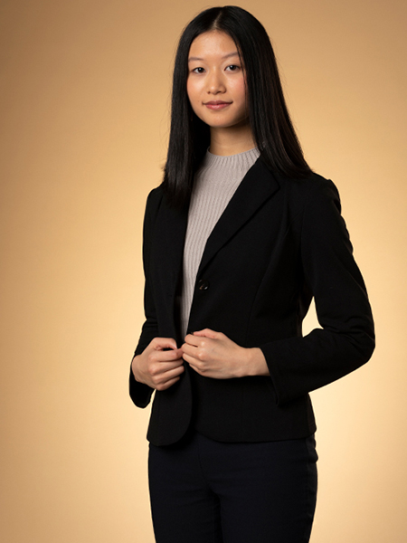 Amanda Ong, wearing a black suit and brown sweater, smiles in front of a brown background. 
