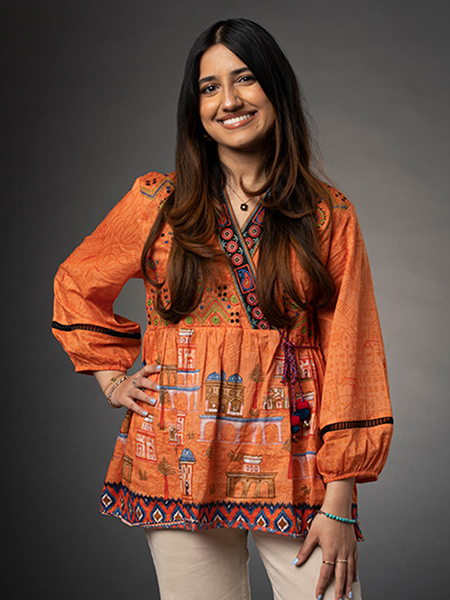 Duaa Zaheer, wearing an orange patterned tunic and tan pants, smiles in front of a gray background. 