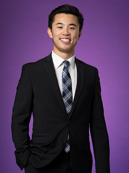 Eric Fan, wearing a black suit jacket, white shirt and dark blue striped tie, smiles for a portrait in front of a purple background. 