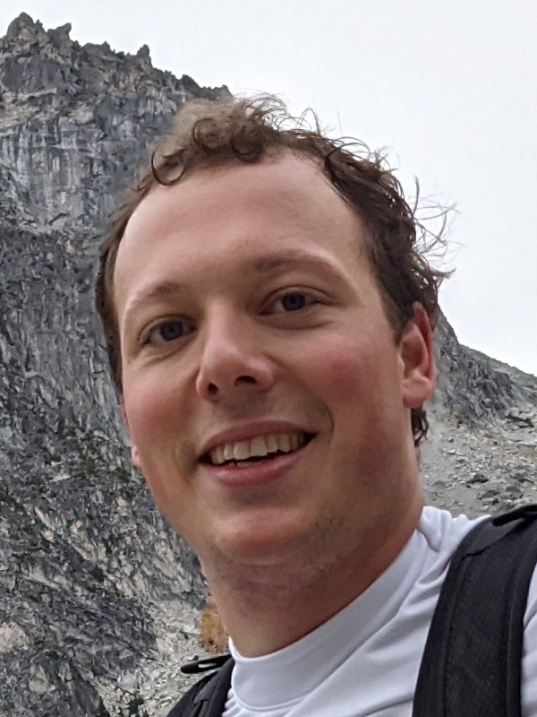 Portrait of Lancelot Wathieu innfront of a rocky peak and overcast sky, smiling and wearing a white technical shirt and black strapped backpack.
