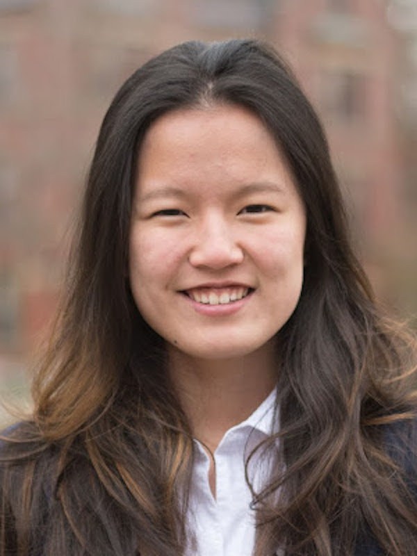 Portrait of Rachel Hong against a blurred backdrop of a brick building, smiling and wearing a white button up cotton blouse and a navy suit jacket.