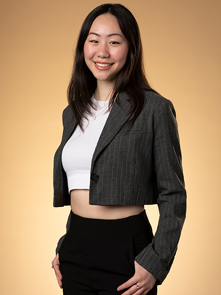 May Wang, wearing a gray striped suit jacket, white shirt and black pants, smiles in front of a brown background.