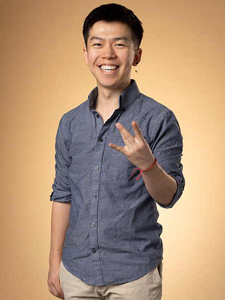Richard Li, wearing a blue shirt and tan pants, smiles in front of a brown background. 