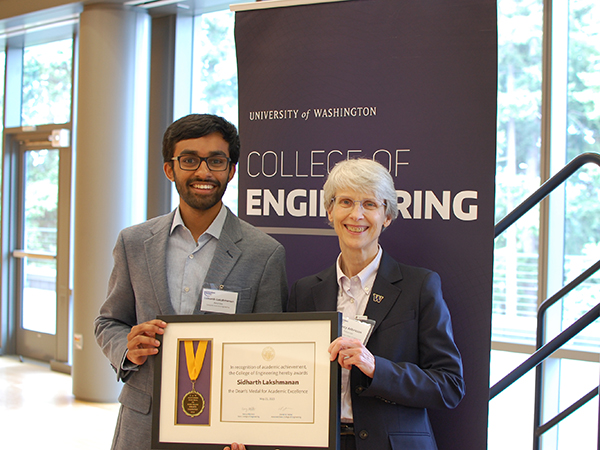 Sidharth Lakshmanan, wearing a blue shirt, glasses and gray suit, smiles next to Nancy Allbritton, wearing a pink shirt and navy suit with a W pin in the lapel. The pair are holding a medal and placard honoring Lakshmanan and are standing in front of a purple University of Washington College of Engineering banner. 