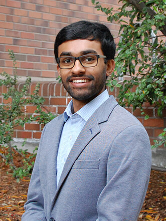 Sidharth Lakshmanan, wearing glasses, a blue shirt and a gray suit jacket, smiles for a portrait in front of a brick building and tree to his left. 
