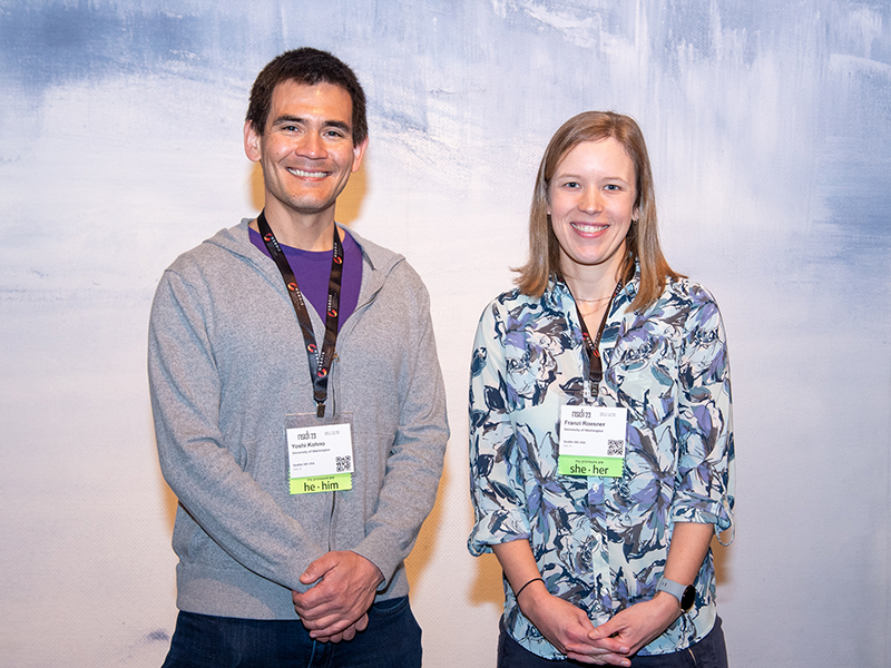 Tadayoshi Kohno and Franziska Roesner smiling and standing side by side, hands clasped in front of them, against a wall painted with visible brush strokes in shades of blue, both wearing lanyards with NSDI name tags around their necks. Kohno is wearing a grey zip-up sweatshirt over a purple t-shirt, and Roesner is wearing a blue floral-patterned blouse with the sleeves rolled up and a smartwatch with a blue wristband.