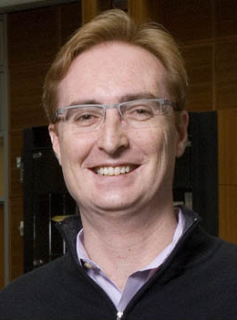 Portrait of David Wetherall against a dark building interior, smiling and wearing wireframe glasses and a black zip-up top over a lavender collared shirt.