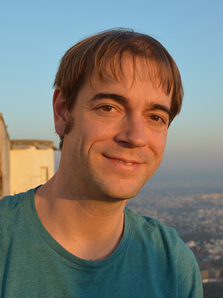 Portrait of Thomas Rothvoss smiling in a blue-green t-shirt with hazy blue sky and part of an old sand-colored building overlooking a city behind him.