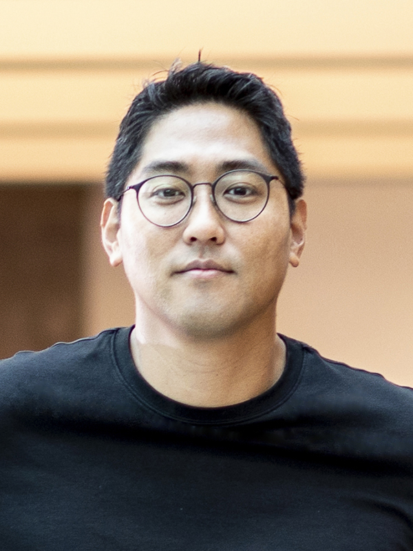 Portrait of Sewoong Oh wearing eyeglasses with thin, round dark frames and a black t-shirt against a warmly lit building interior.