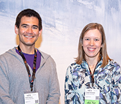 Tadayoshi Kohno and Franziska Roesner smiling and standing side by side against a wall painted with visible brush strokes in shades of blue, both wearing lanyards with NSDI name tags around their necks. Kohno is wearing a grey zip-up sweatshirt over a purple t-shirt, and Roesner is wearing a blue floral-patterned blouse.