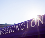The word "Washington" in University of Washington font in white on a purple fabric banner mostly obscuring campus buildings, backed by a pale blue sky and a burst of sunlight above the "o"