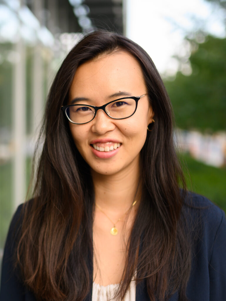 Portrait of Amy Zhang in front of a windowed building exterior and tree-lined boulevard, smiling and wearing black and metallic cat-eye glasses frames, a navy blazer, a cream-colored gathered neckline blouse, gold hoop earrings and a gold pendant necklace.