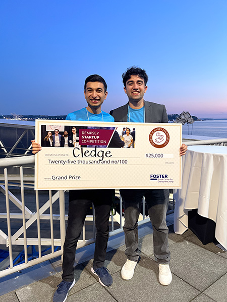 Ayan Gupta, left, and Faraz Qureshi hold a large yellow check bearing the word, "Cledge," representing the grand prize of $25,000 for the Foster Buerk Center for Entrepreneurship Dempsey Startup Competition. Gupta is wearing a blue shirt and black pants and Qureshi is wearing a blue shirt, gray blazer and gray pants. They are standing in front of a railing in front of an evening sunset overlooking the water.