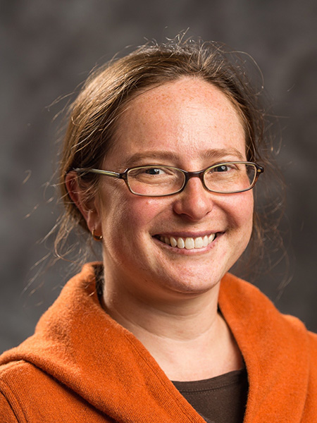 Janet Davis, wearing glasses, brown shirt and orange sweater, smiles for a portrait in front of a brown background. 