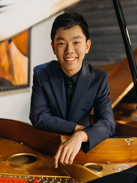 Michael Gu, wearing a black shirt and blue suit jacket, smiles while leaning on a grand piano with the lid open. 