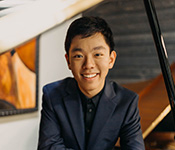 Michael Gu, wearing a black shirt and a blue suit jacket, smiles for a portrait in front of a white and gray wall with a picture frame to his right.