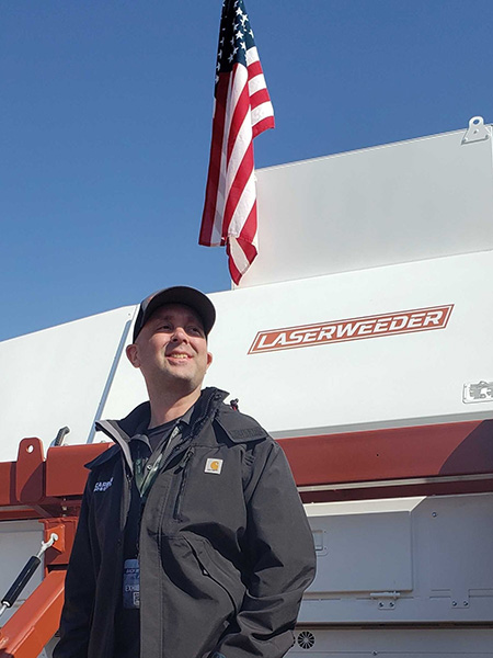 Paul Mikesell, wearing a gray jacket and hat, smiles in front of a white-and-red machine with the words "LASERWEEDER" in a red box across the machine. An American flag is above the machine and Mikesell. 