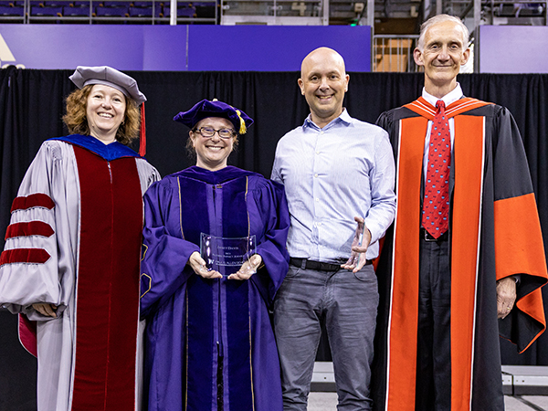 Four people stand in a row smiling on a stage. Three of the four are wearing academic regalia representing their respective Ph.D. alma maters; one is in business-casual wear. The middle two people are holding curved glass plaques.