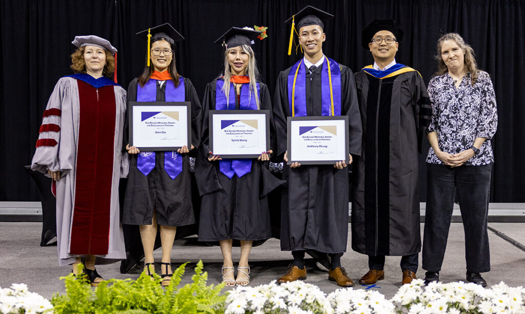 Six people stand next to each other smiling onstage behind a row of white flowers and green foliage. Three of them are wearing graduation regalia consisting of black cap, gold tassel, black gown and purple stole and hold framed award certificates. They are flanked by three people, two wearing the Ph.D. regalia of their alma maters and one dressed in business casual wear.