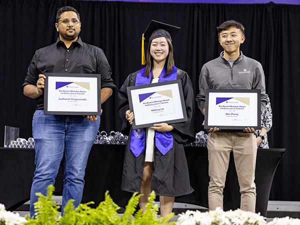 Three people stand smiling onstage holding framed award certificates in front of a table stacked with commemorative diplomas rolled up, and behind a row of white flowers and green foliage. One is wearing business casual wear, one is wearing khaki trousers and an Allen School-branded half-zip pullover, and one is wearing graduation regalia consisting of black cap, gold tassel, black gown and purple stole.