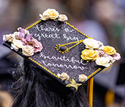Closeup of graduates in regalia from behind, focused on a black undergraduate cap with gold tassel decorated by hand with fabric flowers, gold cord and beads, and glitter and the words "there's a great big beautiful tomorrow" written in script