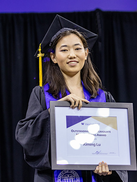A person with long hair wearing black cap with gold tassel, black gown and purple stole smiles holding a framed award certificate.