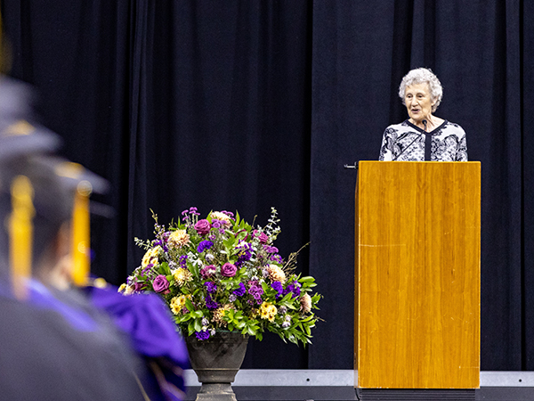 A woman in an ornately patterned white and black blouse stands behind a wood podium with a mic onstage, with a black curtain for a backdrop. There is a large floor vase filled with fresh flowers of varying shades of purple and yellow. The end of several rows are graduates clad in caps and gowns  is visible, blurred, in the far left of the frame.