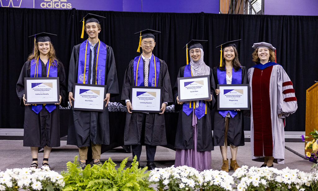 Six people stand next to each other smiling onstage behind a row of white flowers and green ferns. Five of the six are wearing bachelor's graduation regalia of black cap with gold tassel, black gown, and purple stole and hold framed award certificates. The sixth person is wearing Ph.D. regalia from their alma mater.