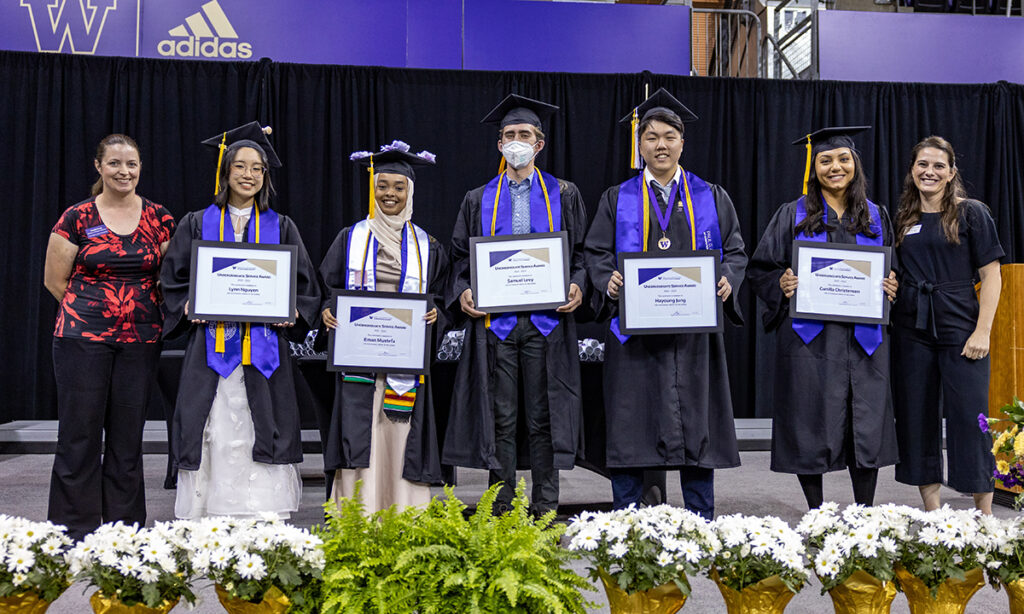 Five graduates standing in a row in regalia inlcuding black cap, gold tassel, black gown and purple stole holding framed award certificates. The winners are flanked by two women on either end in business casual dress. The group is standing onstage behind a row of white flowers and green foliage. The outline of the University of Washington block "W" logo and Adidas logo are visible in gold on a purple hoarding behind the stage.