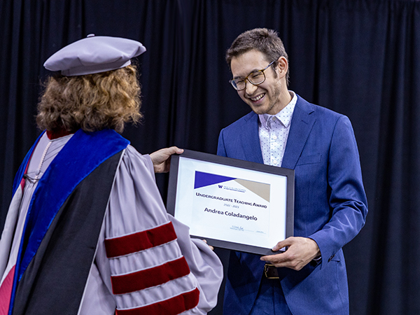 A woman clad in Ph.D. regalia hands a framed award certificate to a man wearing glasses and a vivid blue suit over a pale shirt with matching blue dots.