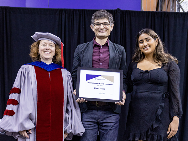 A man wearing glasses, a dark grey suit jacket over a maroon button-down shirt and belted jeans holds a framed award certificate flanked by a woman in Ph.D. regalia and a woman in a black dress with a subtle monochrome dotted pattern and asymmetrical ruffle in front.
