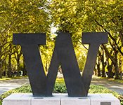 A large, dark "W" stands on a white pedestal in front of a forest background. There is sunlight coming through the trees.